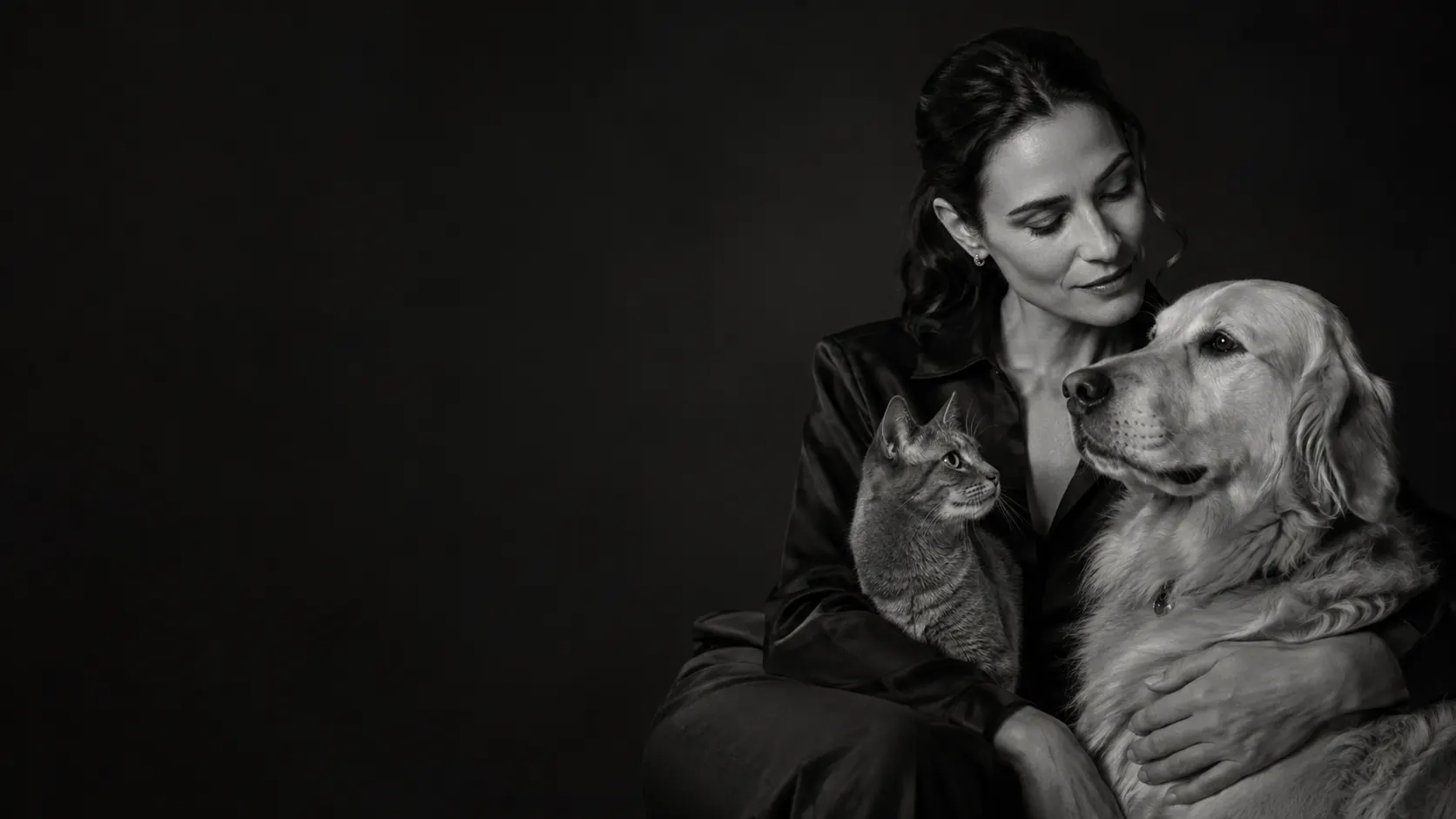 A woman seated in low light with a tabby cat against her chest and a golden retriever resting under her hand, photographed in black and white.